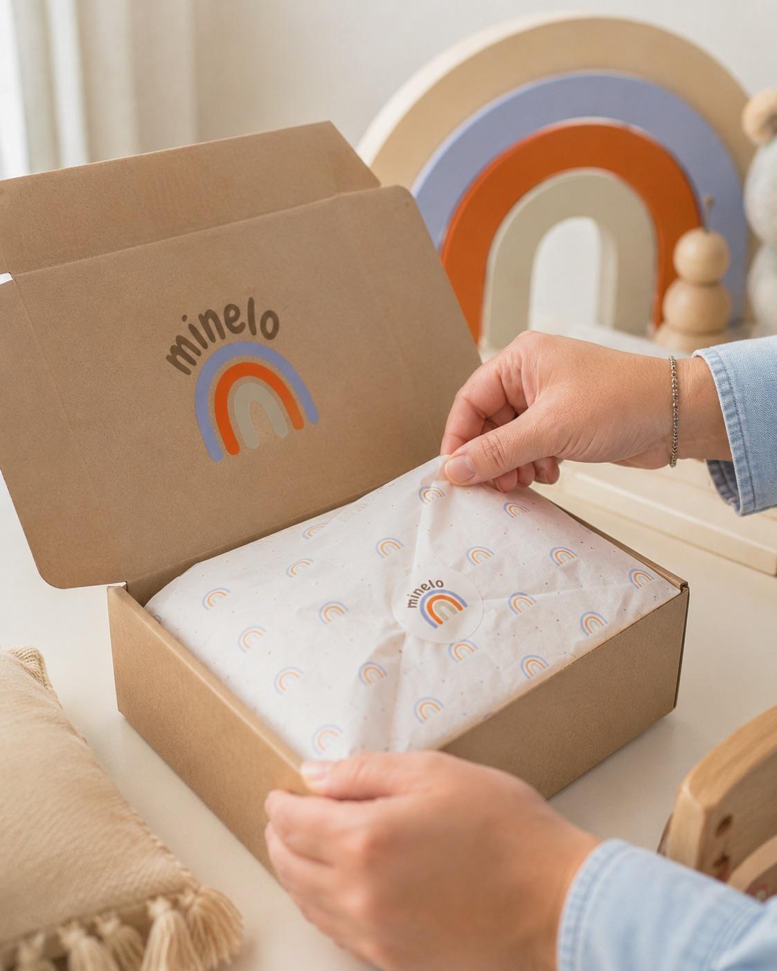 Hände öffnen eine Versandbox mit Minelo-Logo, darin in Seidenpapier eingeschlagene Materialien; im Hintergrund ein Holzregenbogen.