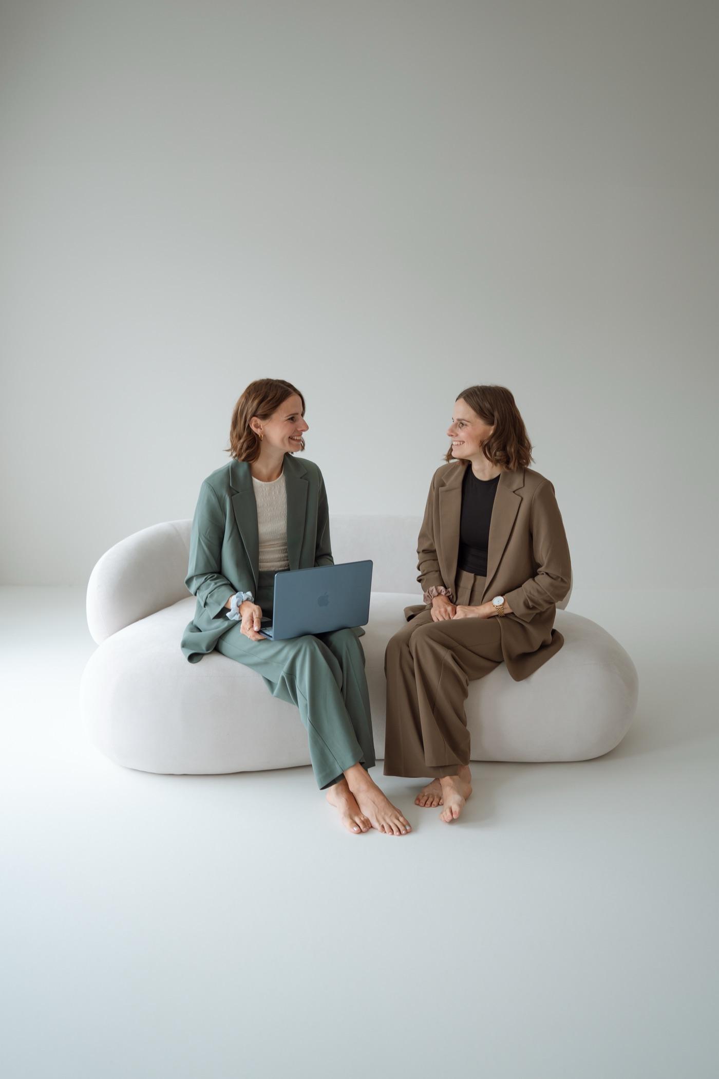 Die beiden Gründerinnen von Minelo sitzen auf einem hellen Sofa und lächeln sich an; eine hält einen Laptop auf dem Schoß.